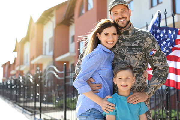 Hombre en uniforme de combate de pie frente a una vivienda con una mujer y un niño pequeño.