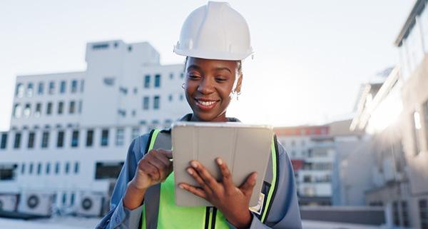 A woman wearing a hard hat and safety vest looks at a tablet.