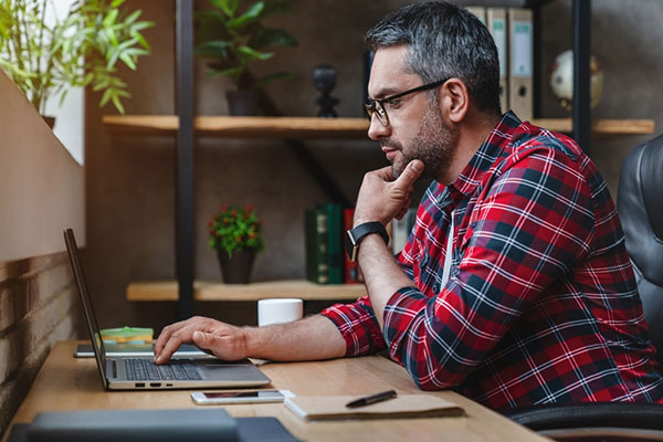 A man in a plaid flannel shirt looks at a laptop while sitting in an office.